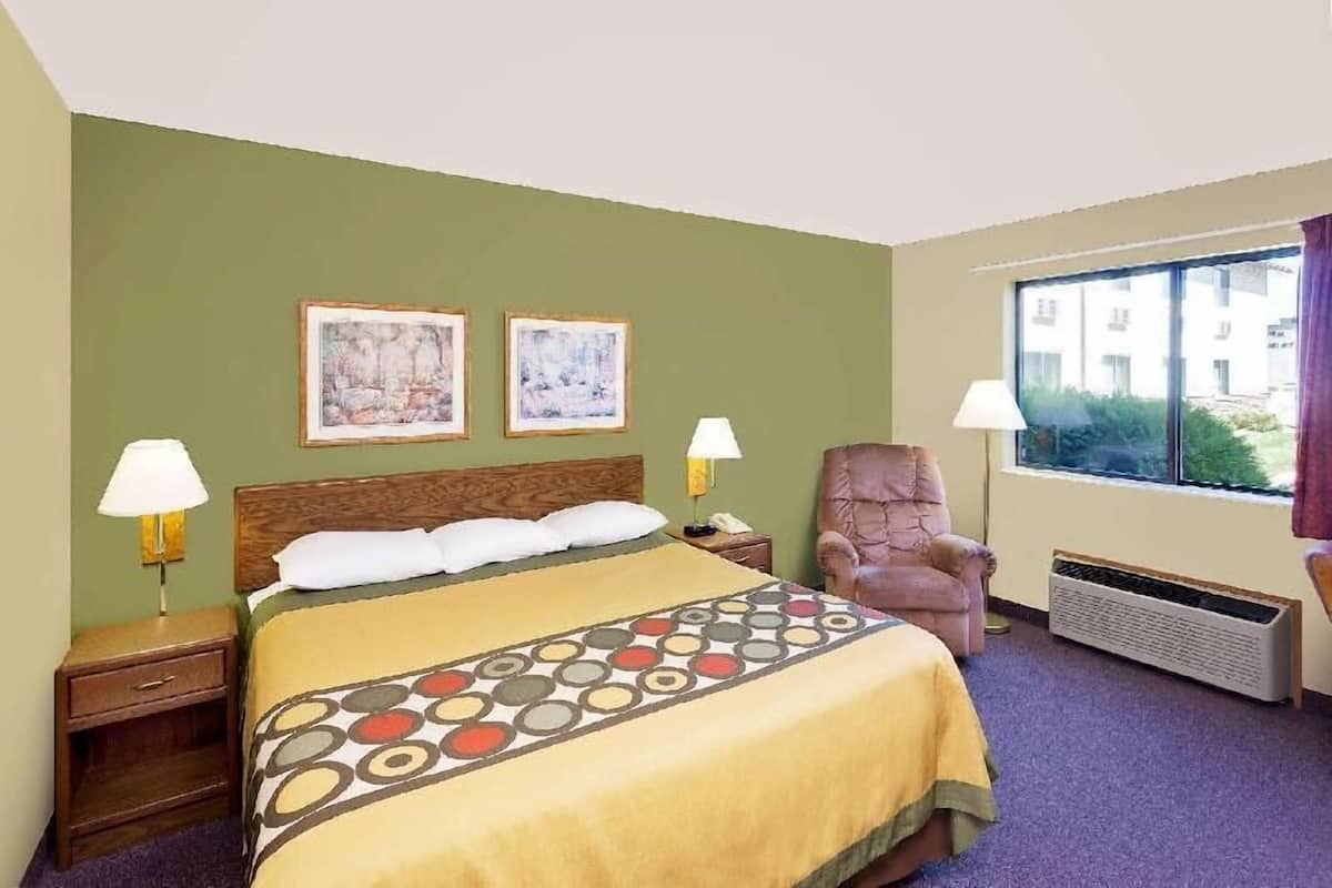 Une chambre d’hôtel avec un lit, deux lampes de chevet, une chaise, une fenêtre donnant sur un paysage agréable et deux tableaux encadrés accrochés au mur.