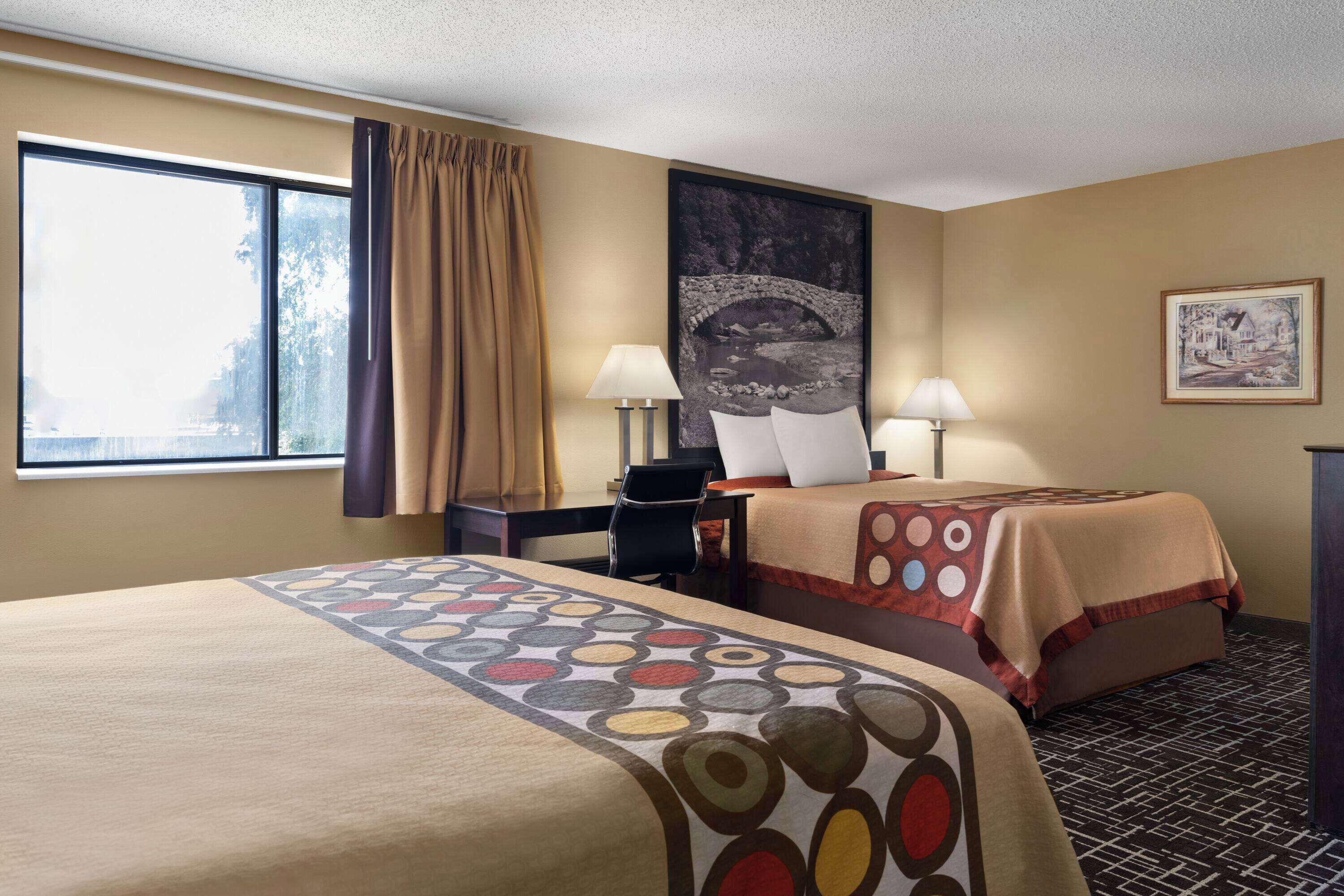 A hotel room with two beds, a desk, two lamps, a large window with curtains, and a framed picture on the wall.