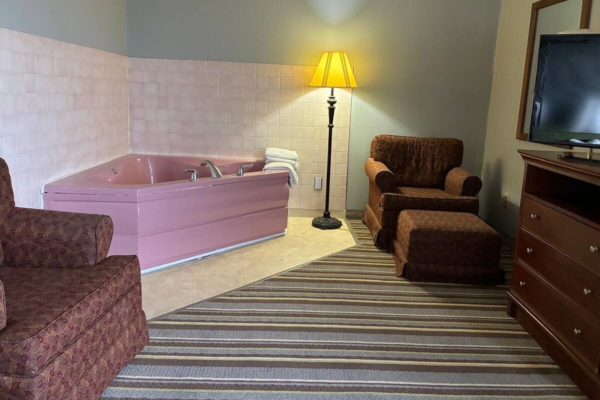 A bathroom with a pink bathtub, two chairs, a floor lamp, and a television on a wooden cabinet.