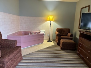 Un baño con una bañera rosa, dos sillas, una lámpara de pie y un televisor sobre un mueble de madera.