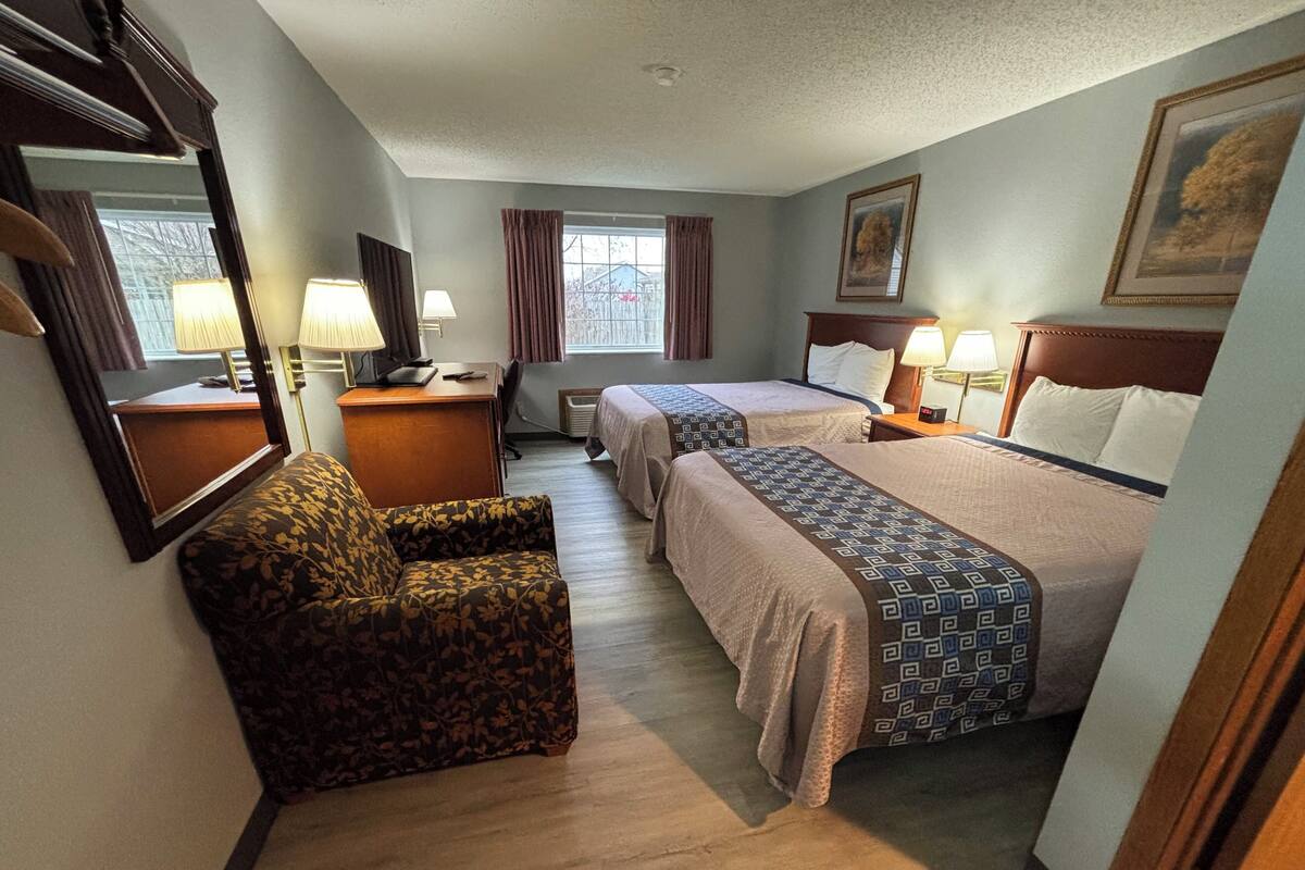 A hotel room with two beds, a desk, a chair, a window with curtains, and a painting on the wall.