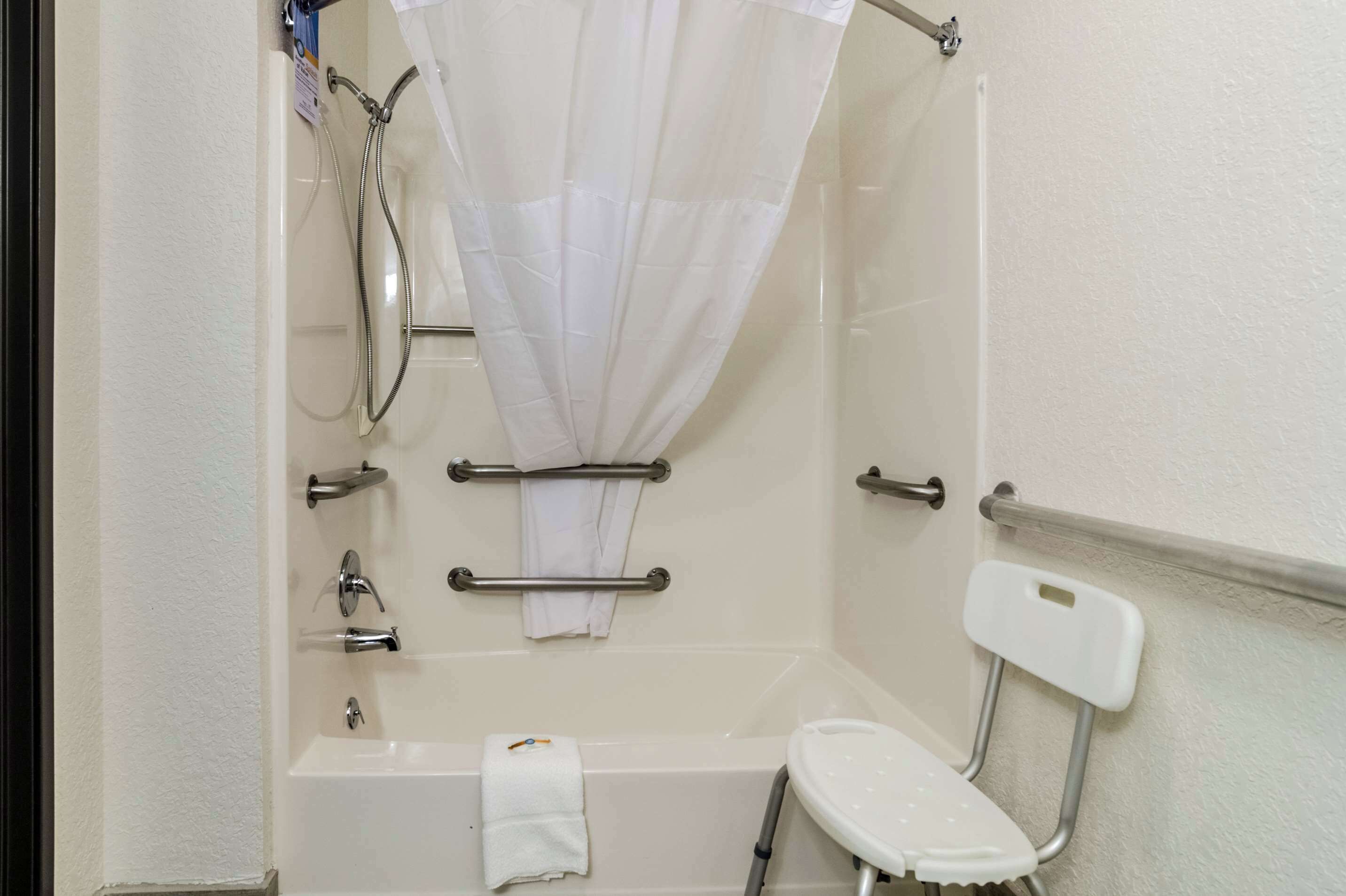 Un baño con bañera, ducha, barras de apoyo, una silla blanca y un perchero para toallas.