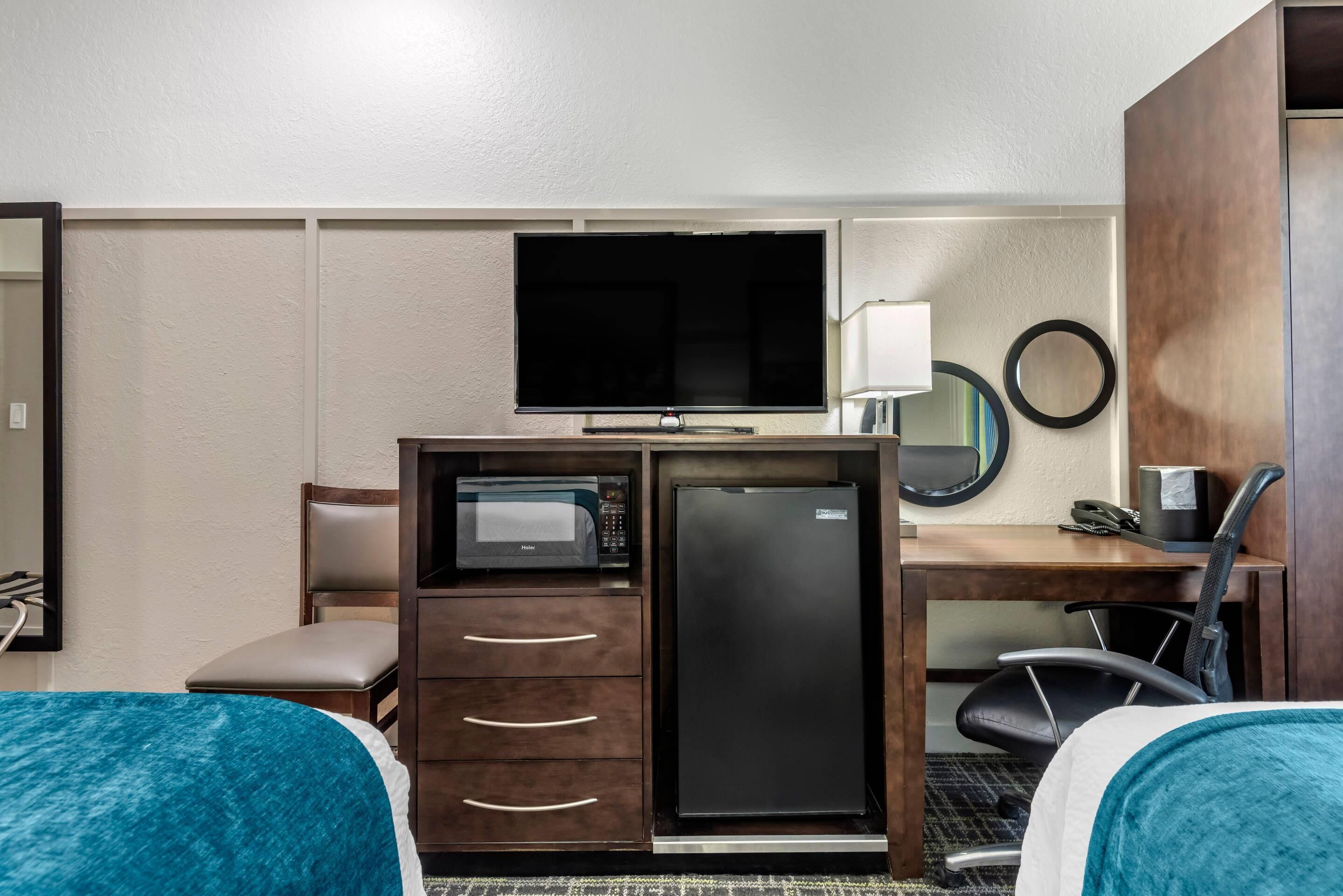 A hotel room with a bed, a desk with a computer, a microwave, a refrigerator, a chair, a mirror, and a television.