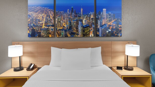 A hotel room with a large cityscape painting above the bed, two bedside tables with lamps, and a wooden headboard. - Days Inn by Wyndham Waukegan (Waukegan)
