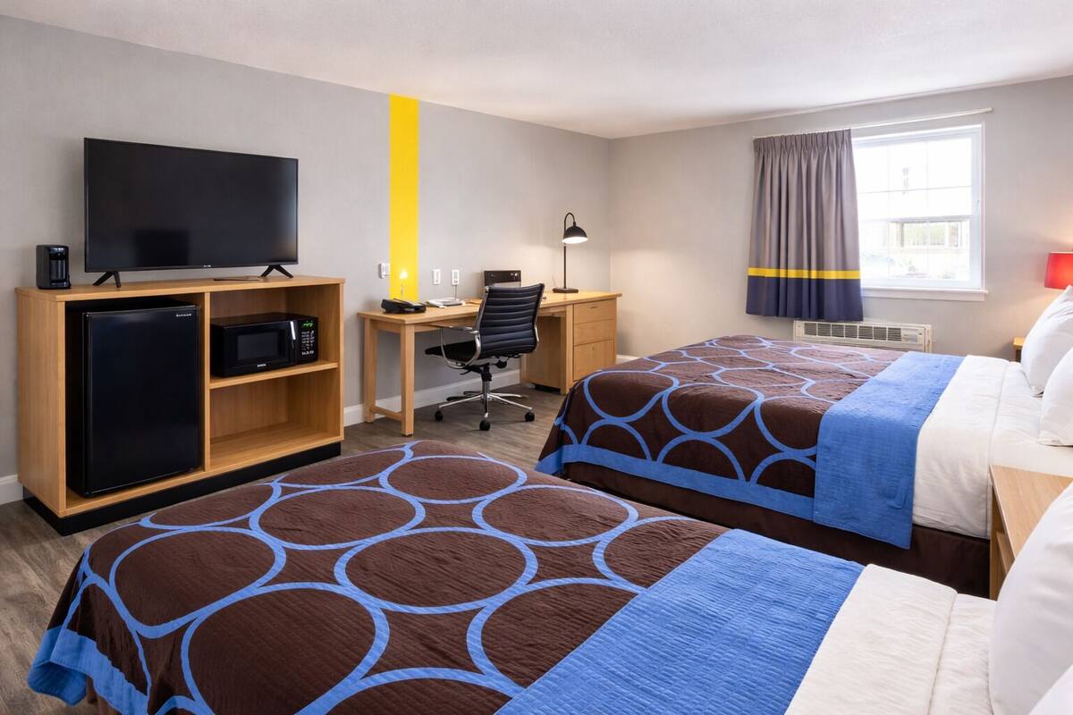 A hotel room with two beds, a desk, a television, a refrigerator, and a window with curtains.