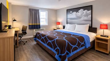 Habitación de hotel con cama, escritorio, silla, dos lámparas de noche, ventana con cortinas, aire acondicionado y un cuadro grande en la pared.