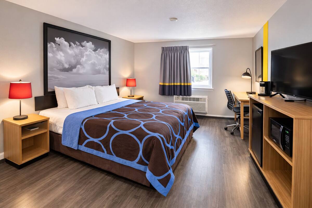 A hotel room with a bed, bedside tables, a TV, a desk, and a large window with curtains.