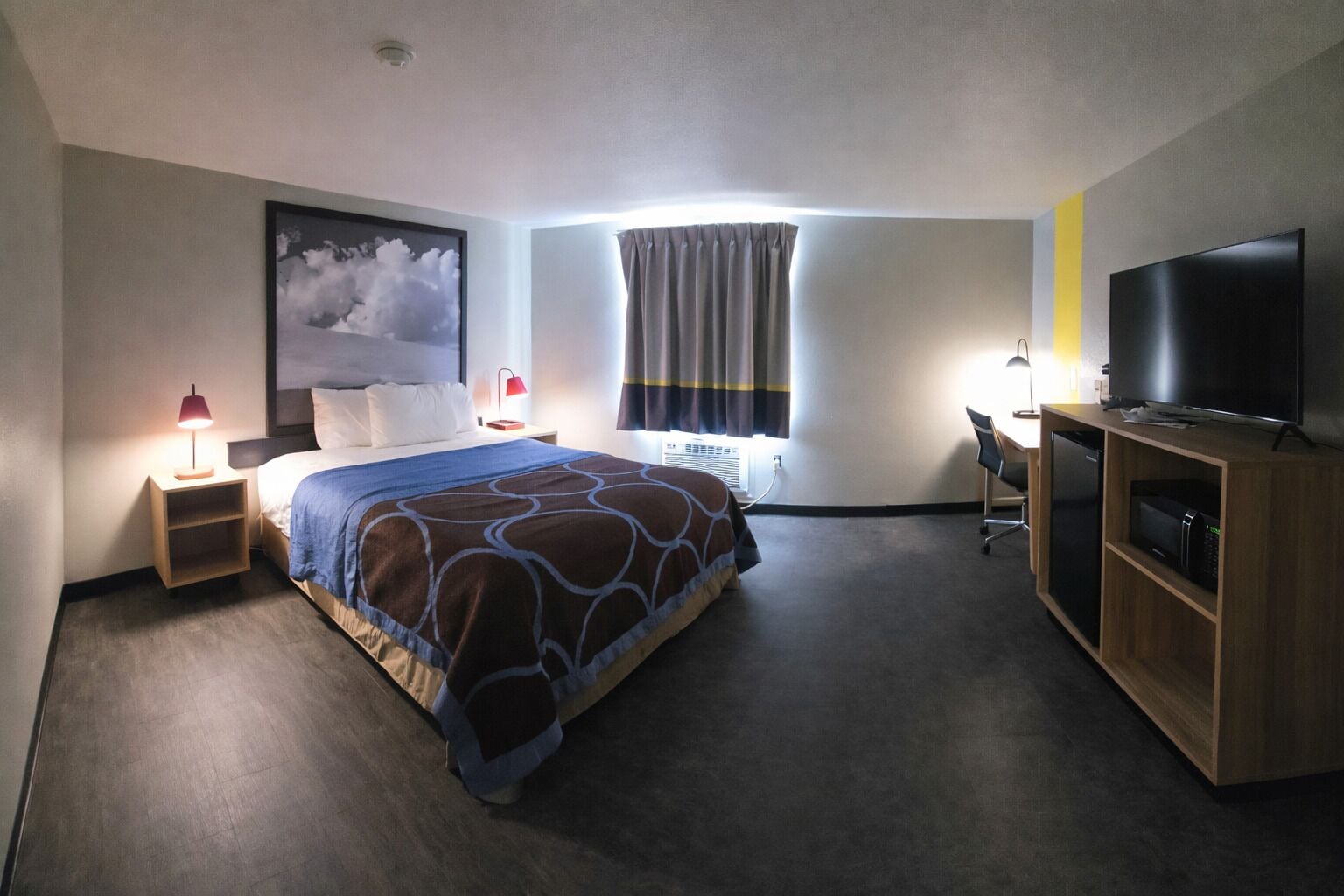 A hotel room with a bed, a TV, a desk with a chair, a window with curtains, and a wall-mounted TV.