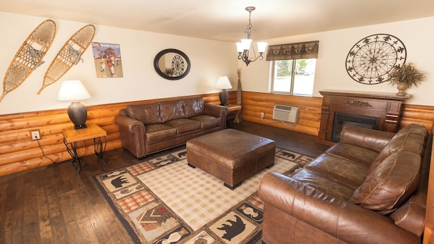 L’intérieur d’une cabane en rondins, avec un canapé en cuir marron, un pouf assorti, une table en bois sur laquelle se trouve une lampe, une cheminée et une grande fenêtre.