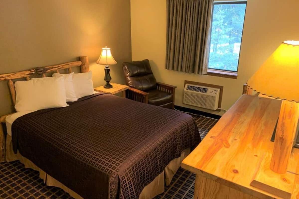 A hotel room with a large bed, a wooden nightstand, a chair, a desk, and an air conditioning unit.
