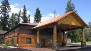 Un bâtiment de style chalet en rondins, doté d’un porche couvert, de piliers en pierre et offrant une vue dégagée sur la forêt environnante.