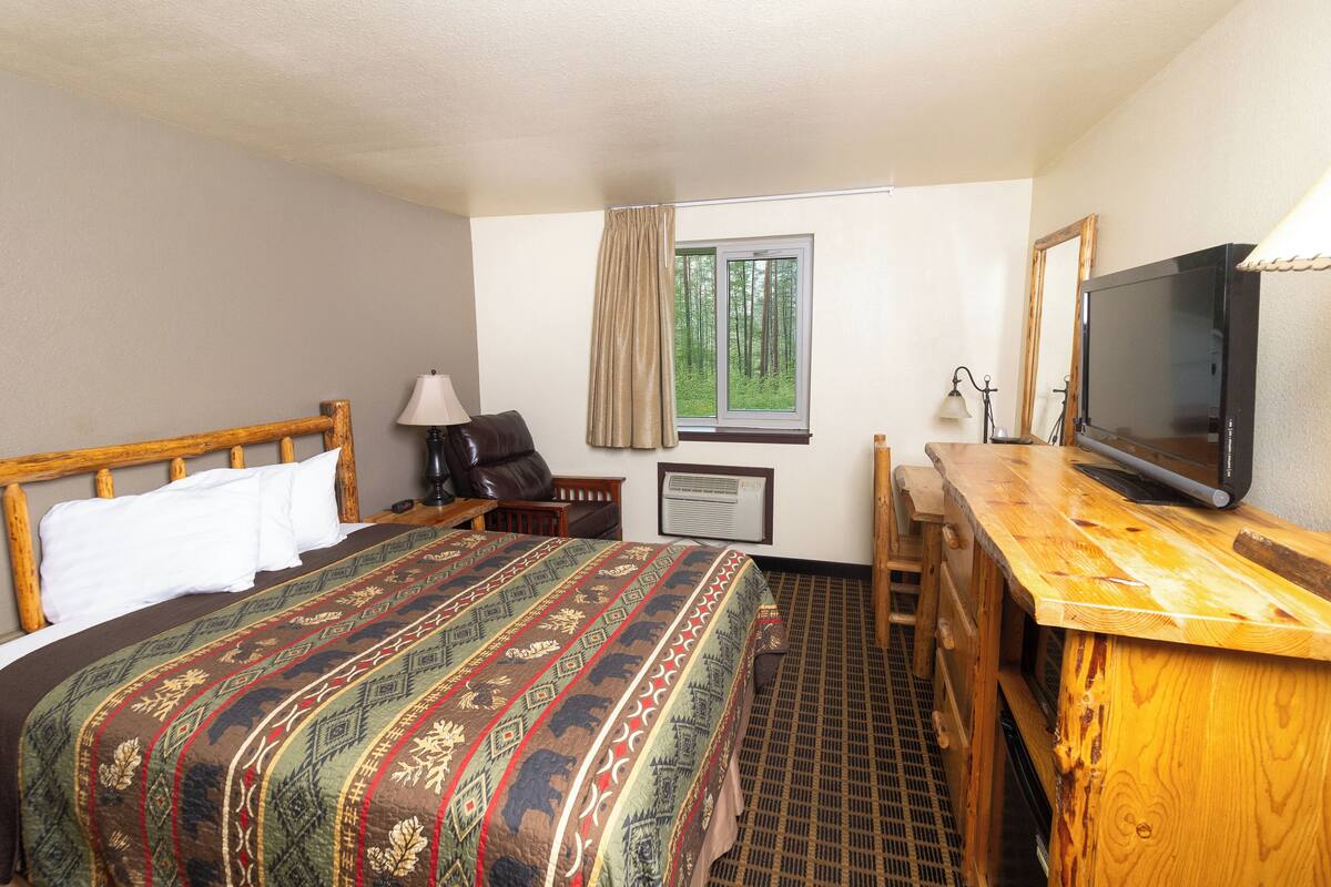 A hotel room with a large bed, a wooden desk, a television, and a window with a view of trees.