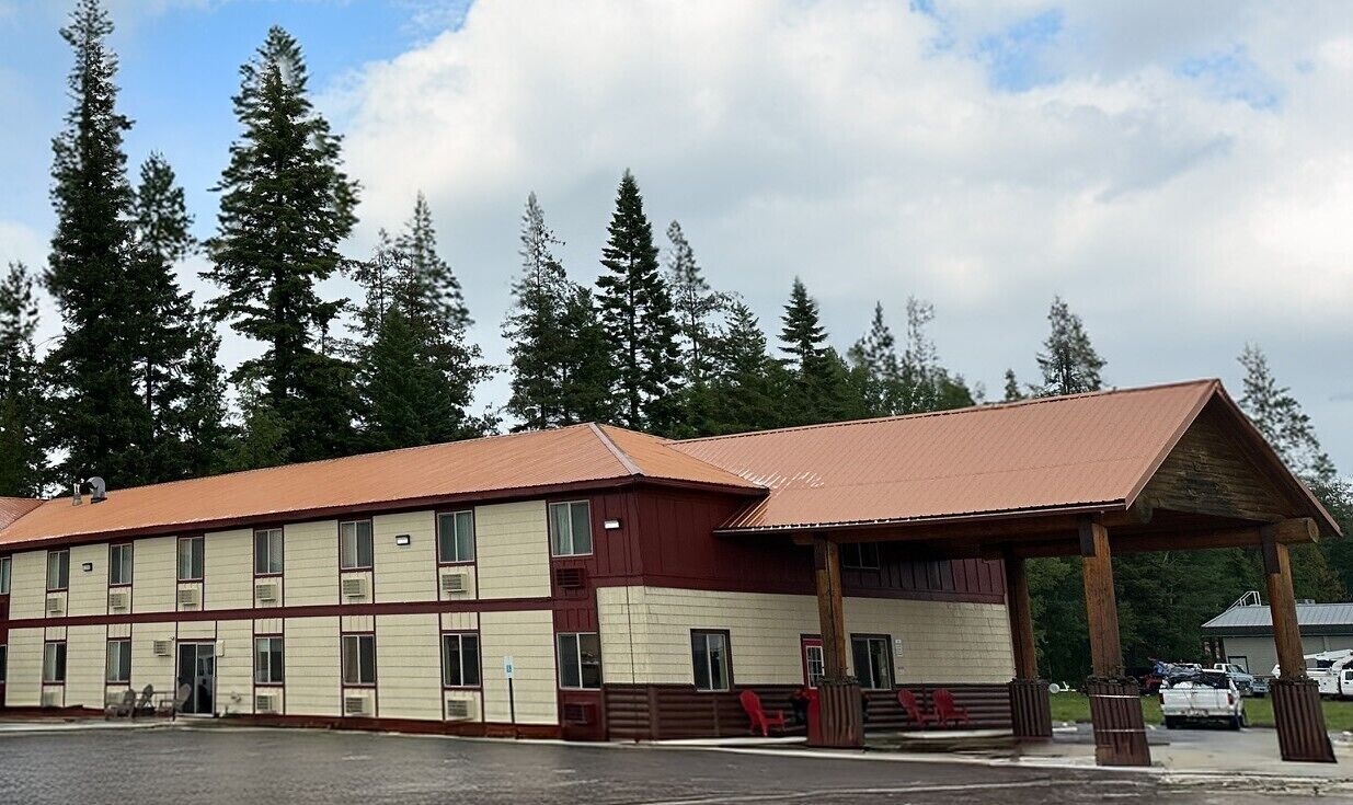 A two-story motel with a red roof and beige walls, featuring a covered parking area and a small adjacent building.