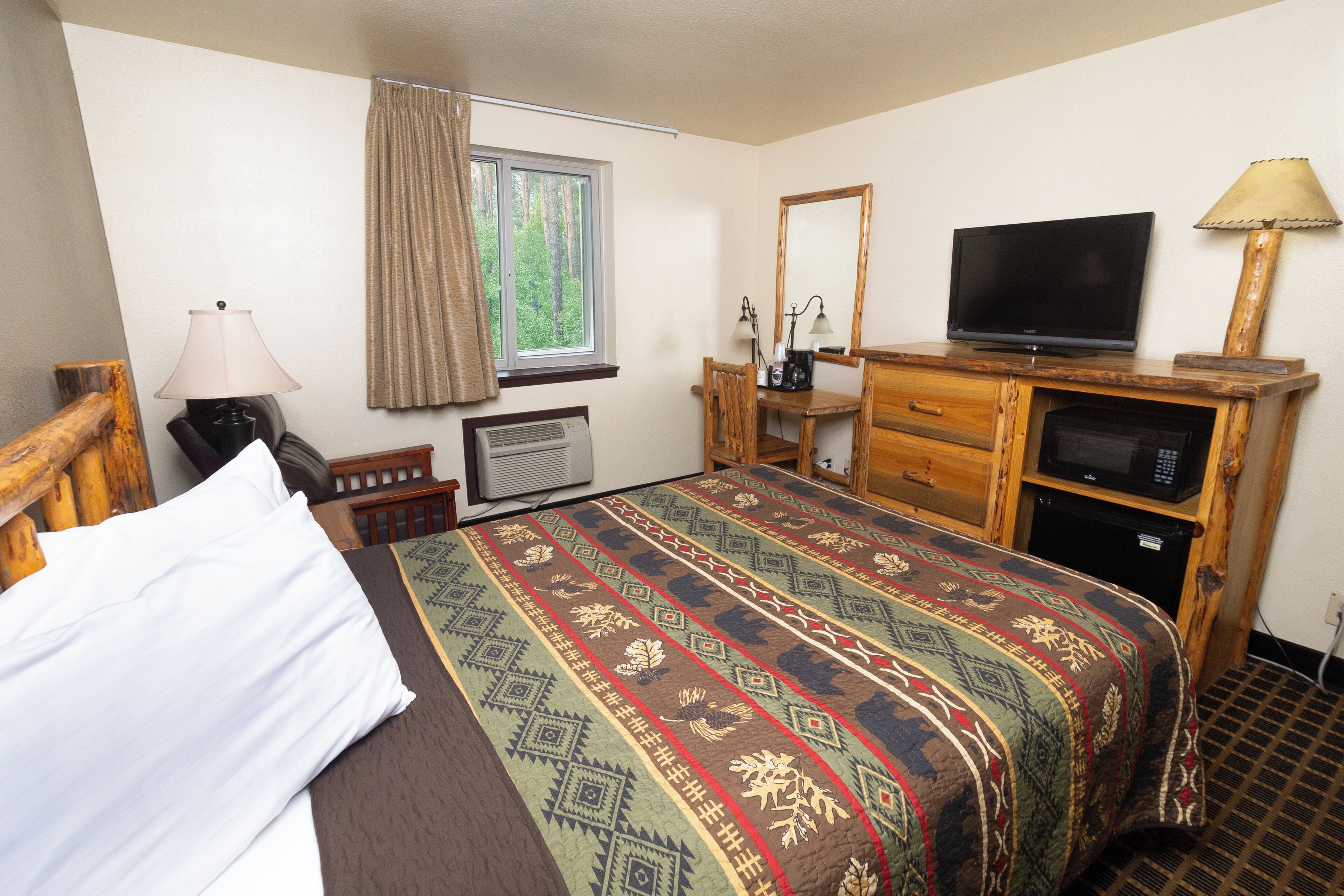 A hotel room with a bed, a television, a desk with a computer, a mirror, and a window with curtains.