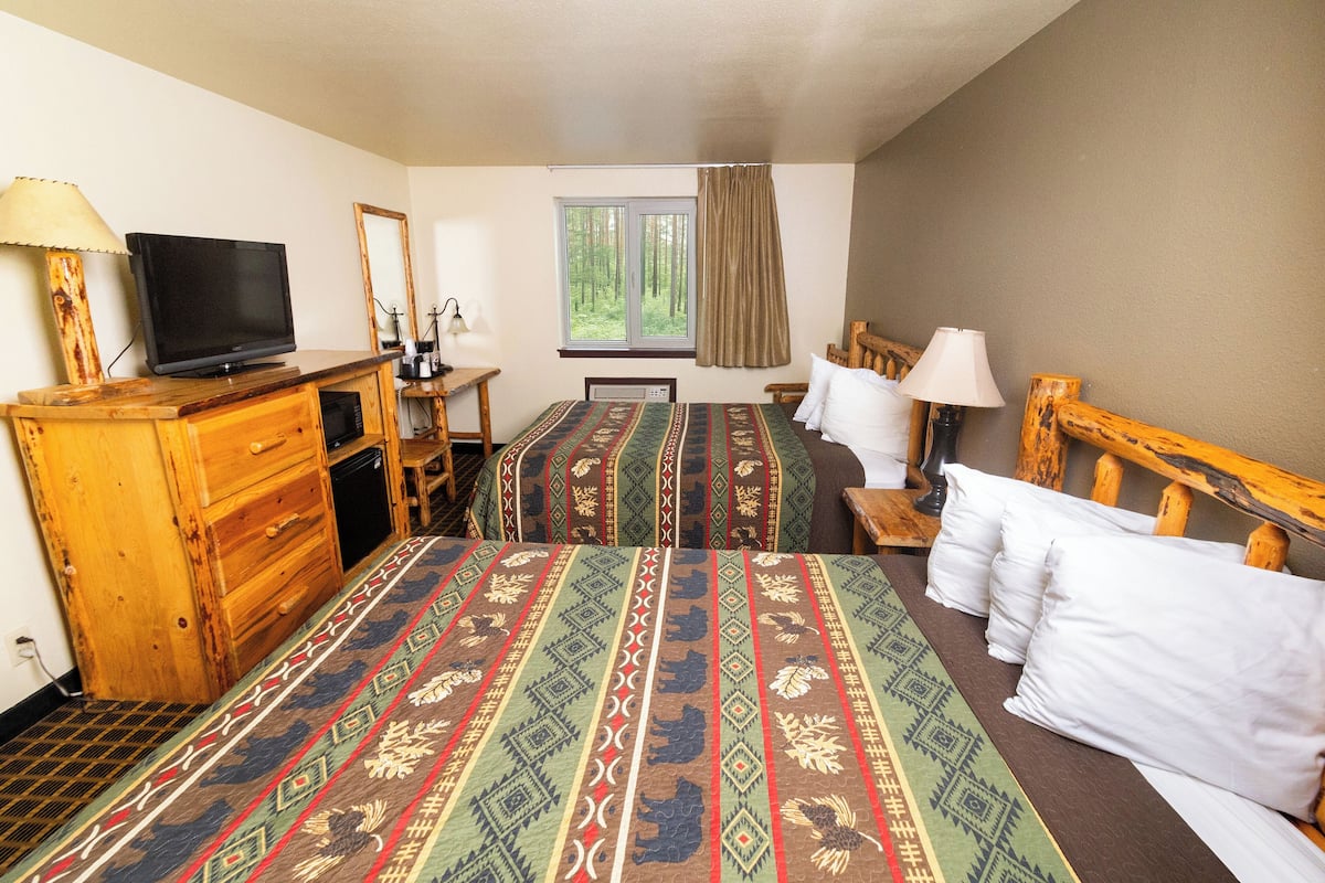 A hotel room with two beds, a TV, wooden furniture, a window with a view of trees, and a desk with a lamp.
