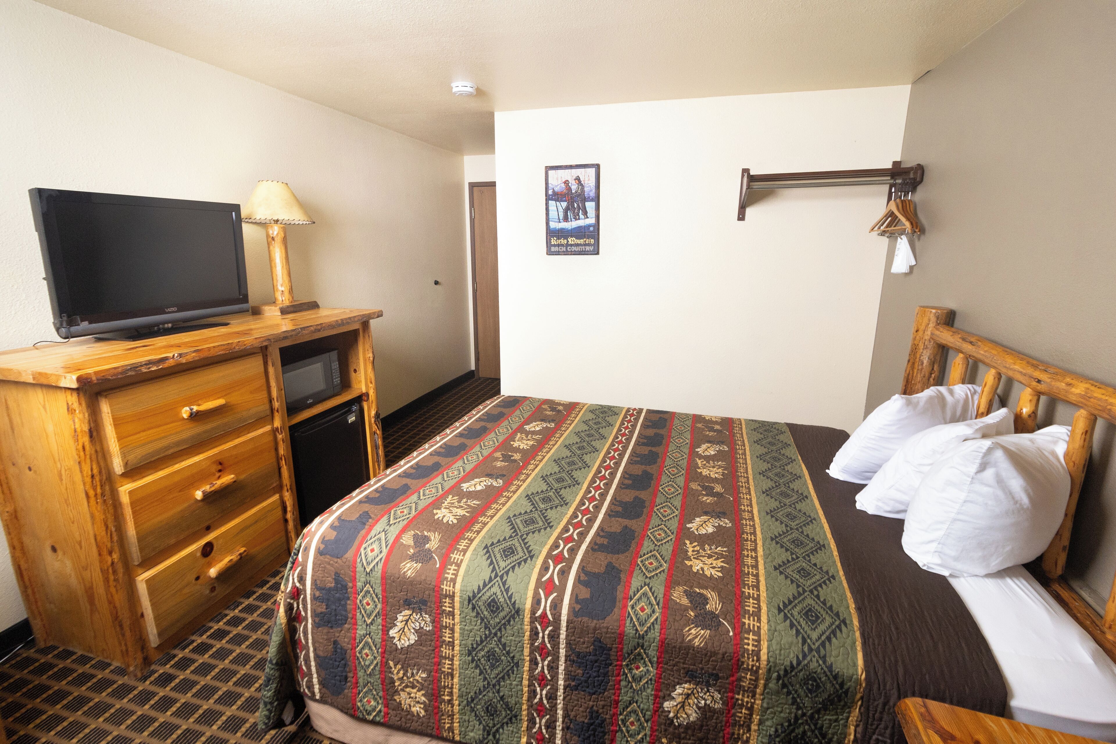 A hotel room with a bed, a TV on a wooden dresser, a small desk, and a wall-mounted coat rack.