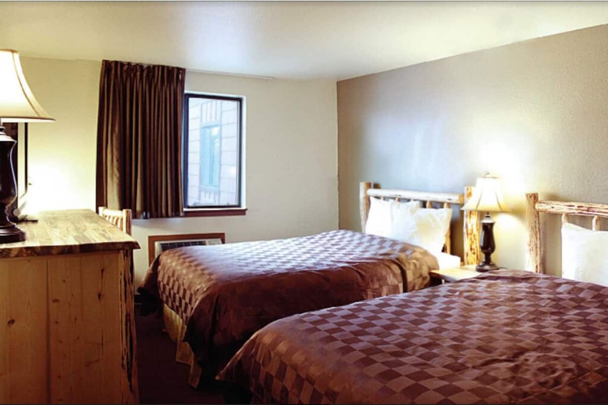 A hotel room with two beds, a wooden desk, a window with curtains, and a nightstand with a lamp.