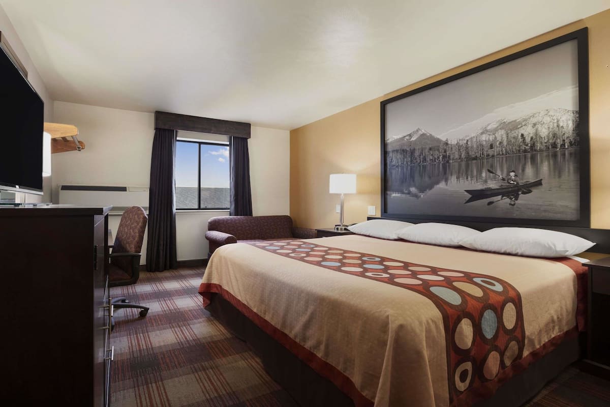 Une chambre d’hôtel avec un grand lit, un bureau, une chaise, une fenêtre avec des rideaux, une lampe et un tableau représentant un lac et des montagnes.