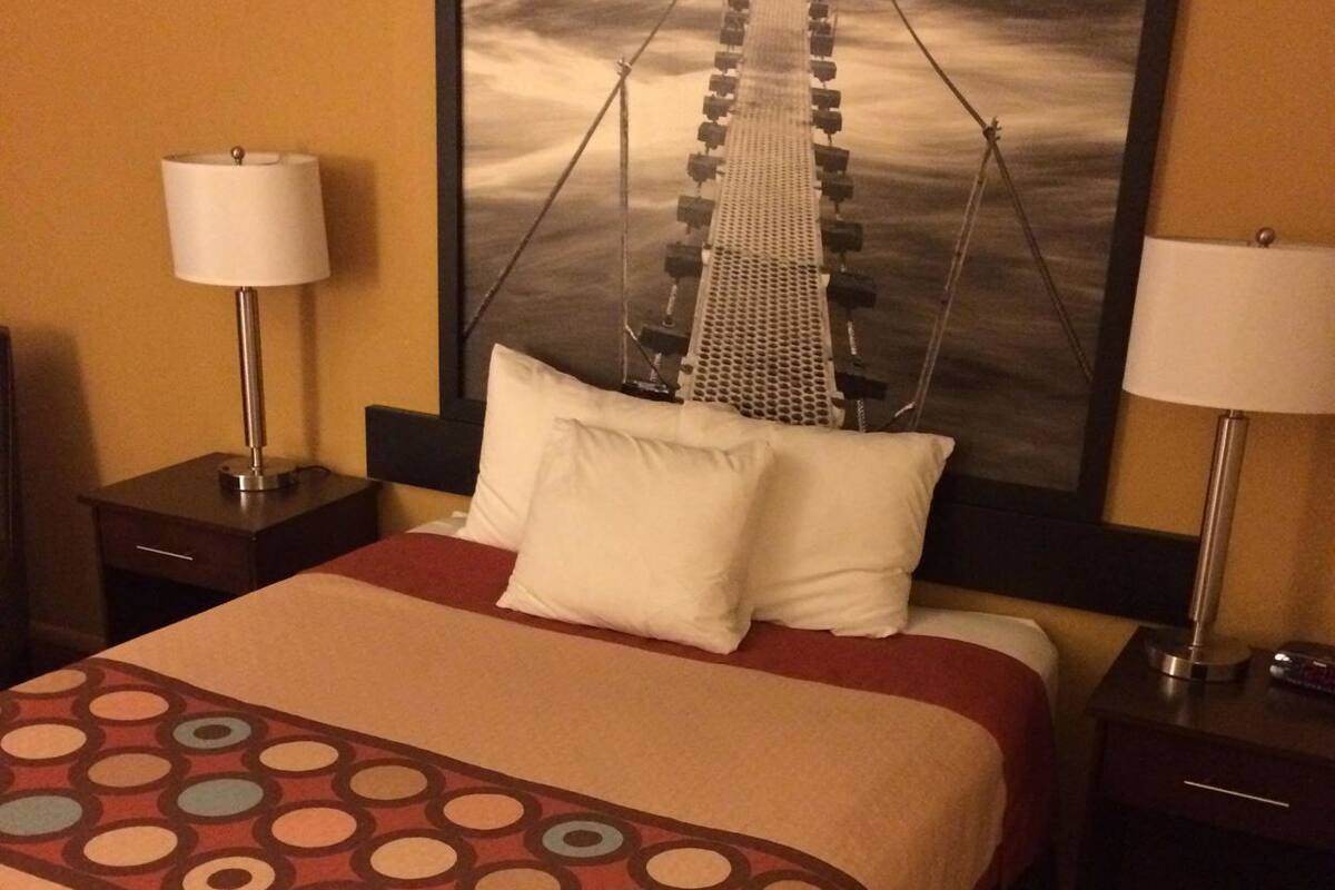Une chambre d’hôtel avec un lit, deux tables de chevet, des lampes, un grand tableau encadré représentant un pont et une couvre-lit à motifs.