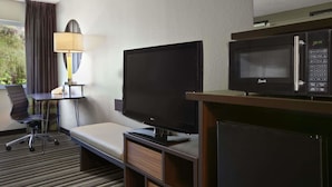A hotel room with a flat-screen TV, a microwave, a desk with a lamp, a chair, and a bed.