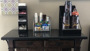 A wooden bar counter with storage drawers, a coffee station with disposable cups and a coffee machine, and a shelf with snacks.