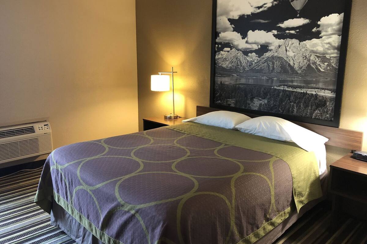 Een hotelkamer met een bed, een nachtkastje, een lamp, een groot gepoft schilderij en een raam.