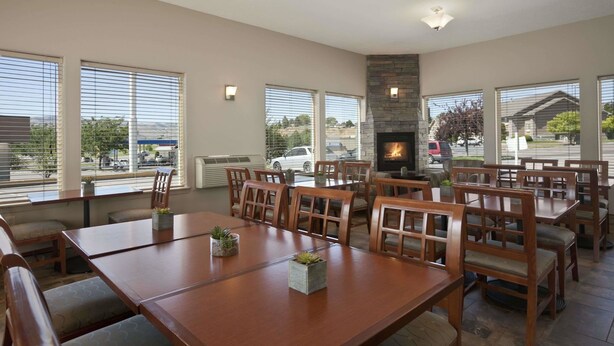 A spacious dining area with multiple wooden tables and chairs, large windows with blinds, and a stone fireplace.