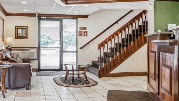 A hotel lobby with a reception desk, a staircase, a seating area with a glass table, and a large window providing a view of the outdoors. - Quality Inn South (Indianapolis)