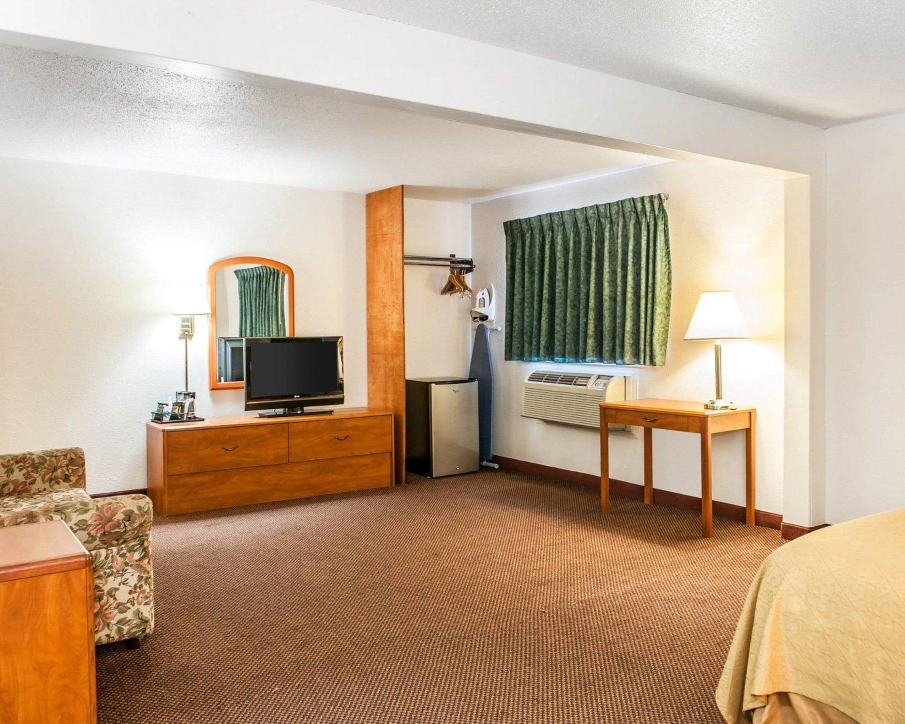 A hotel room with a wooden dresser, a television, a small table with a lamp, a floral-patterned chair, and a window with curtains.