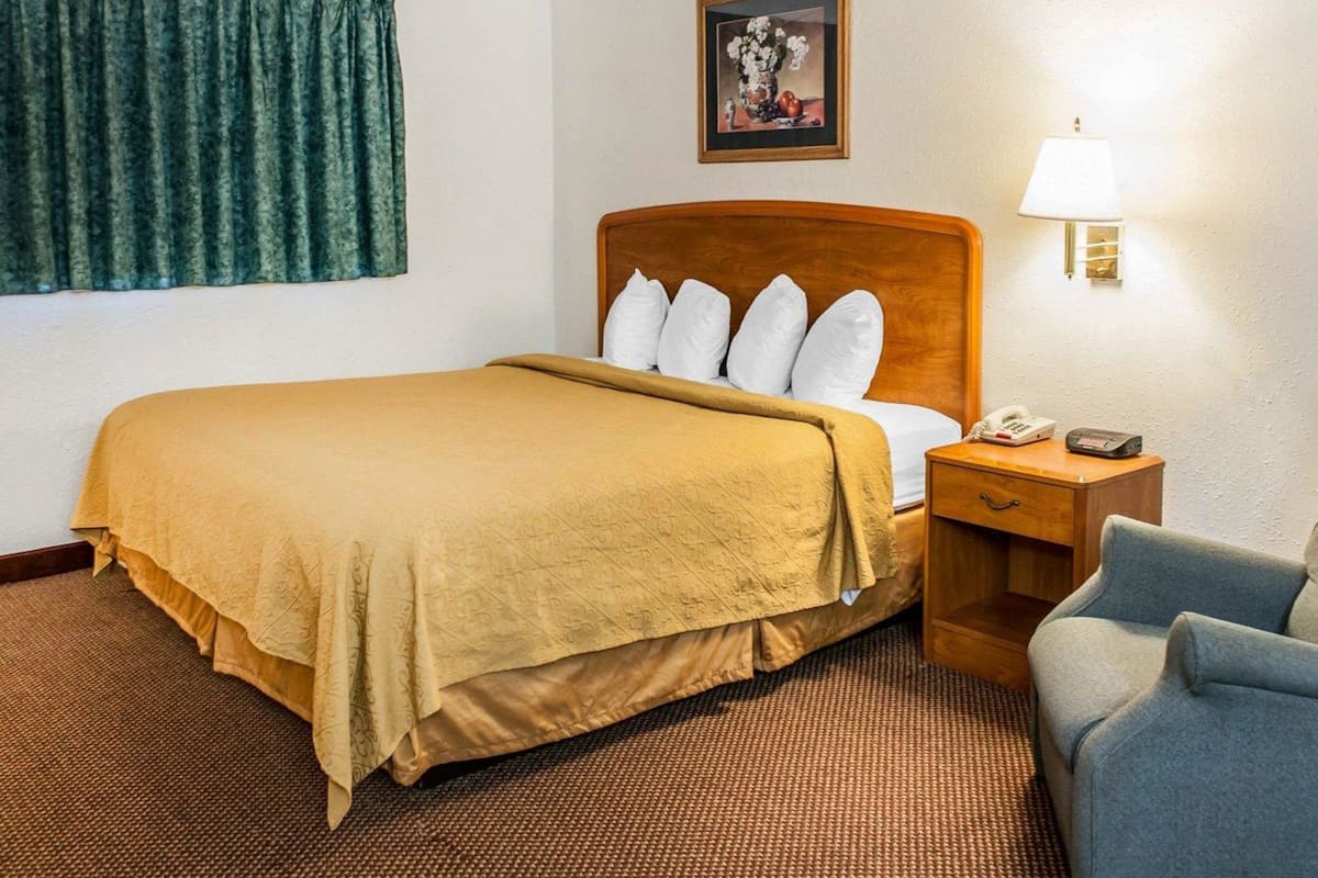 Habitación de hotel con cama, mesita de noche, lámpara, silla y un cuadro en la pared.
