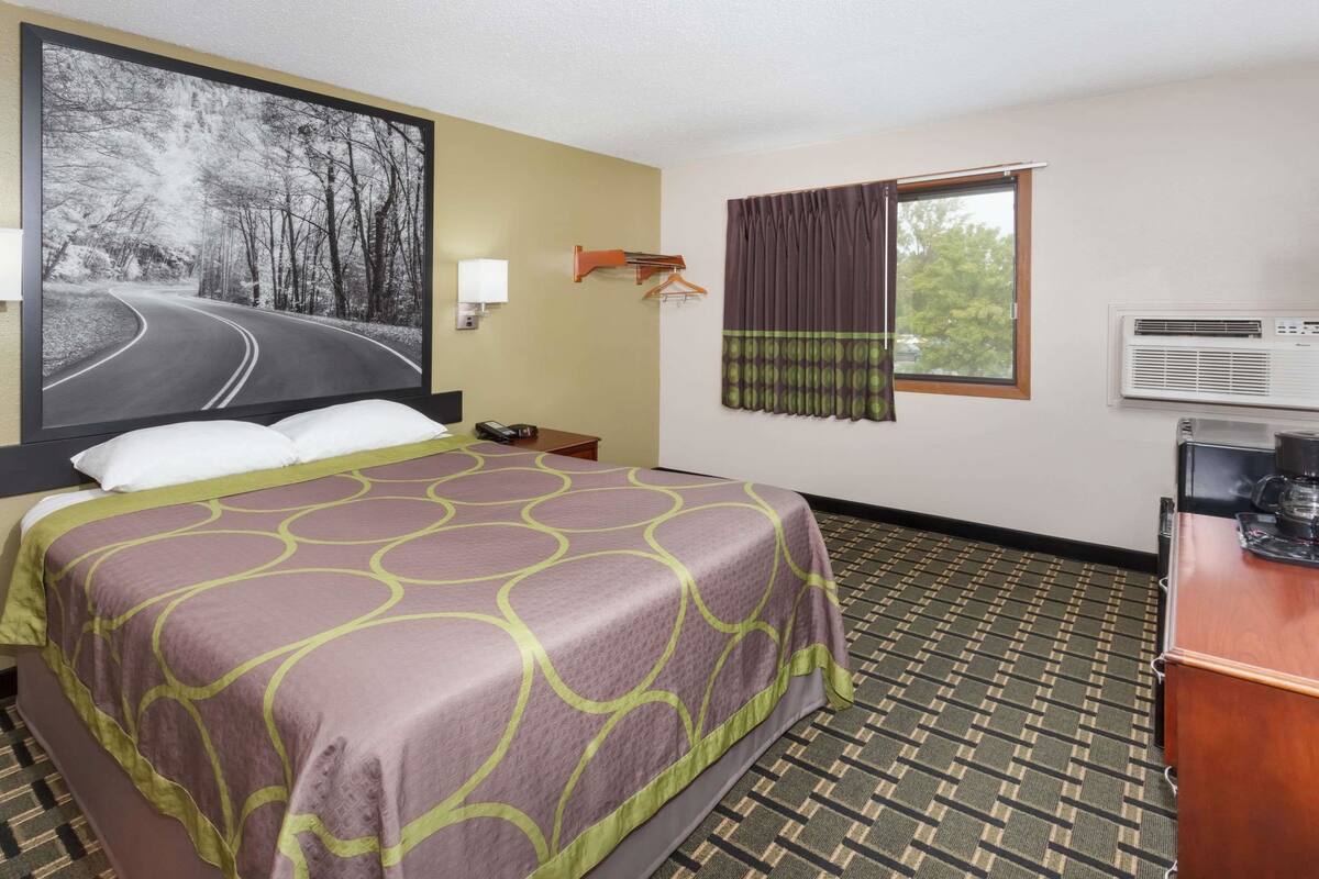 Ein Hotelzimmer mit einem Bett, einem großen Gemälde einer Straße, einer Klimaanlage und einem Fenster mit Vorhängen.
