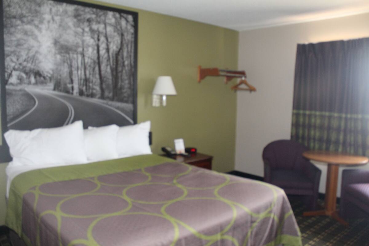 Ein Hotelzimmer mit einem Bett, einem Nachttisch, einer Lampe, einem Sessel, einem Tisch und einem Bild an der Wand.