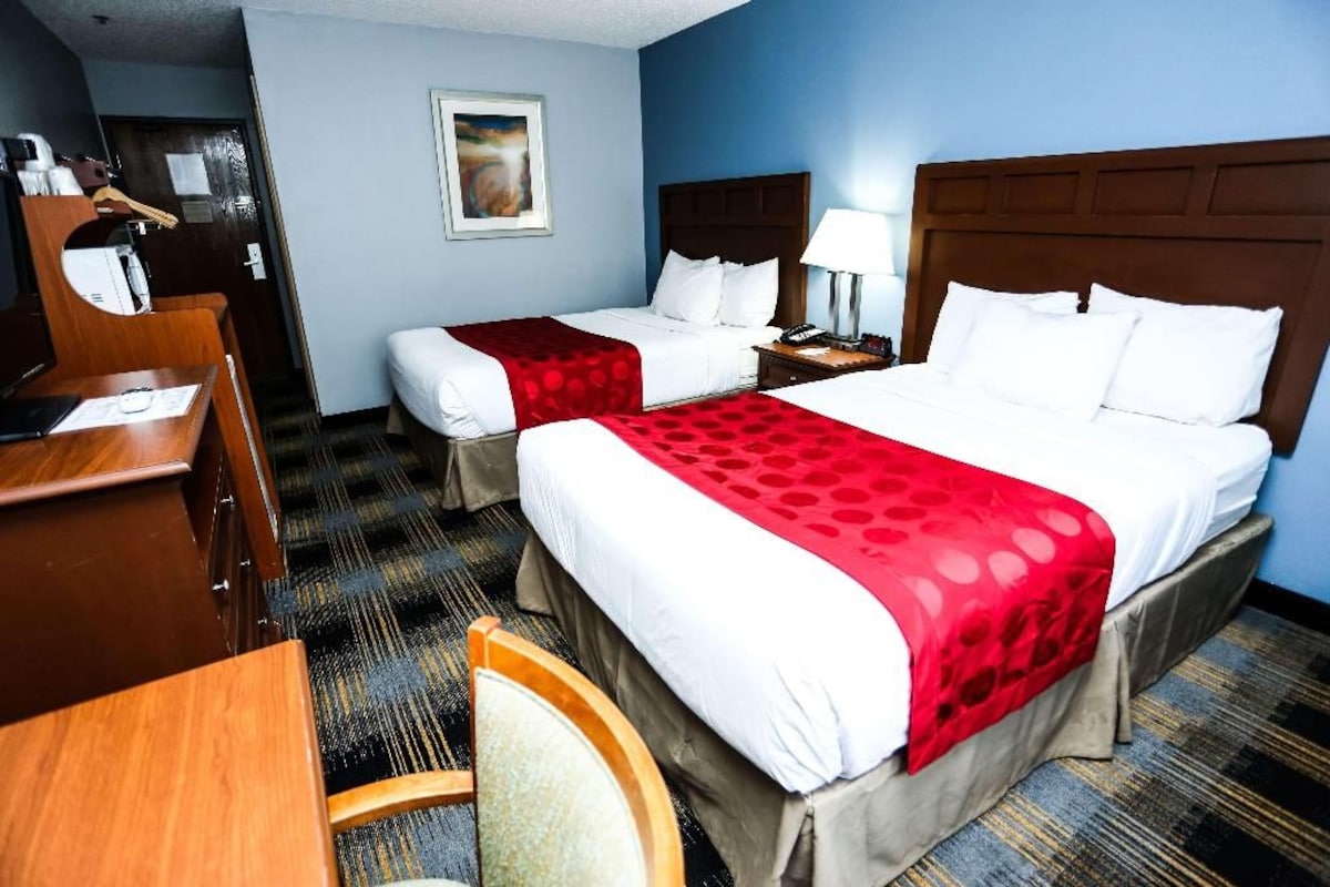 A hotel room with two beds, a desk, a chair, a nightstand, and a painting on the wall.