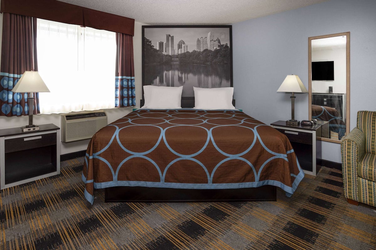 A hotel room with a bed, two bedside tables, a desk, a chair, a large window with curtains, a cityscape painting, and a mirror.