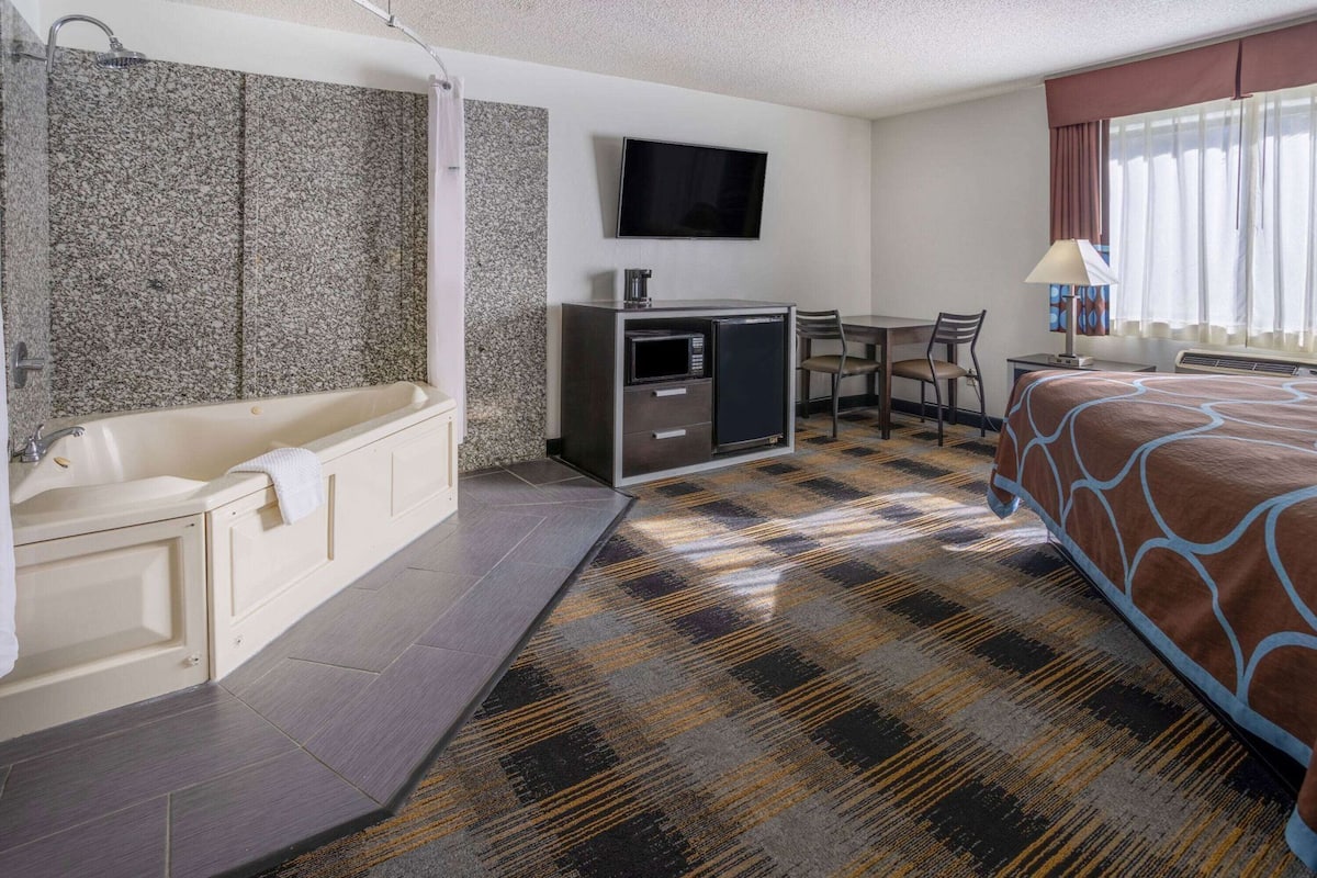 A hotel room with a bathtub, a desk with a flat-screen TV, a bed, and a patterned carpet.