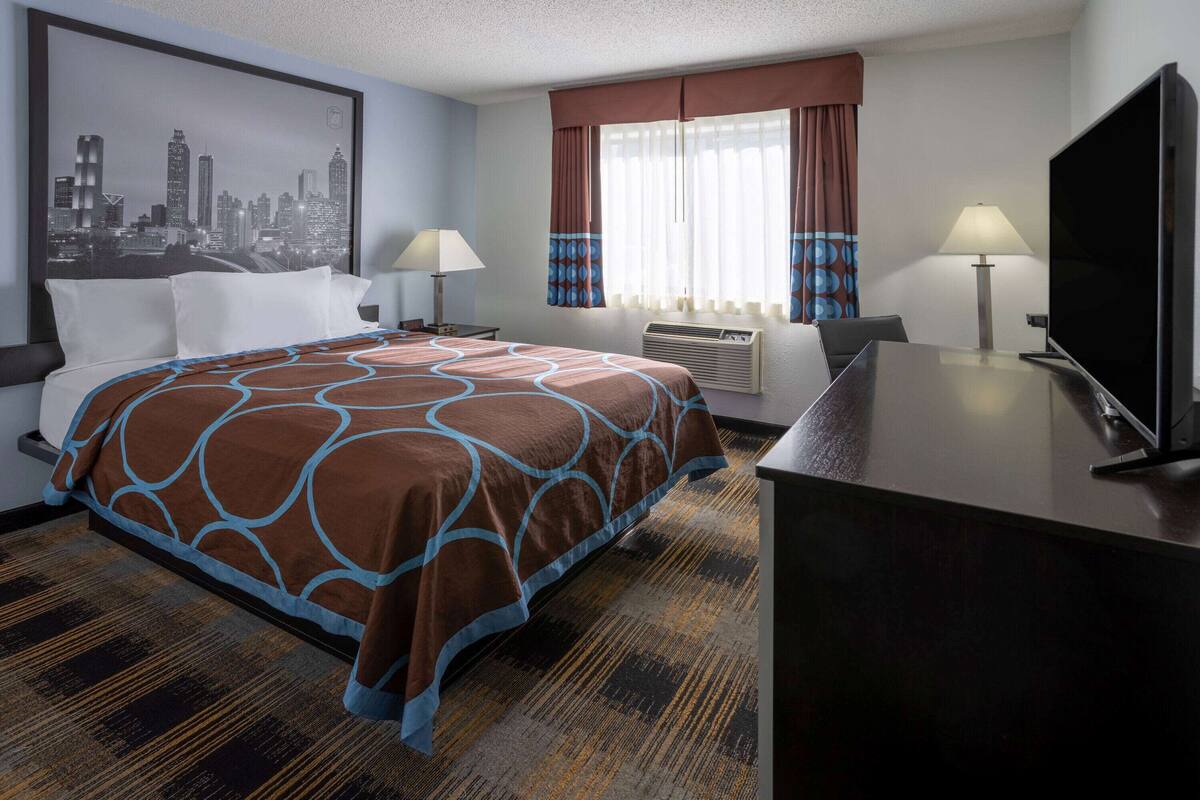 A hotel room with a bed, two bedside tables with lamps, a flat-screen TV, a large window with curtains, and a cityscape painting on the wall.