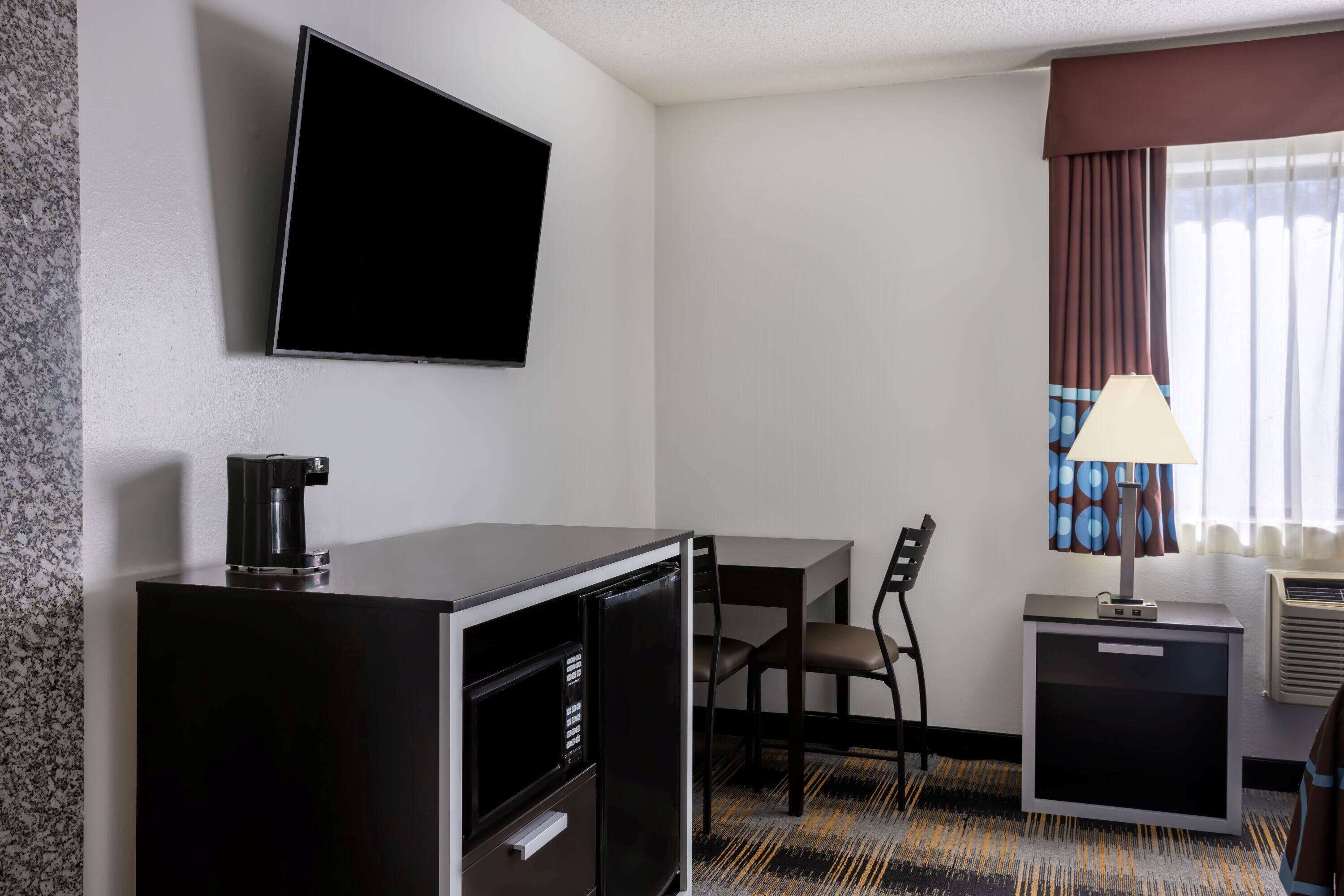 A hotel room with a flat-screen TV mounted on the wall, a small desk with a chair, a coffee machine, a lamp, and a heater.