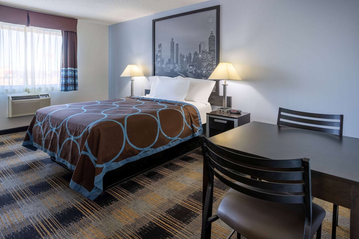 A hotel room with a bed, two bedside lamps, a cityscape artwork on the wall, a dining table with a chair, and a patterned carpet.