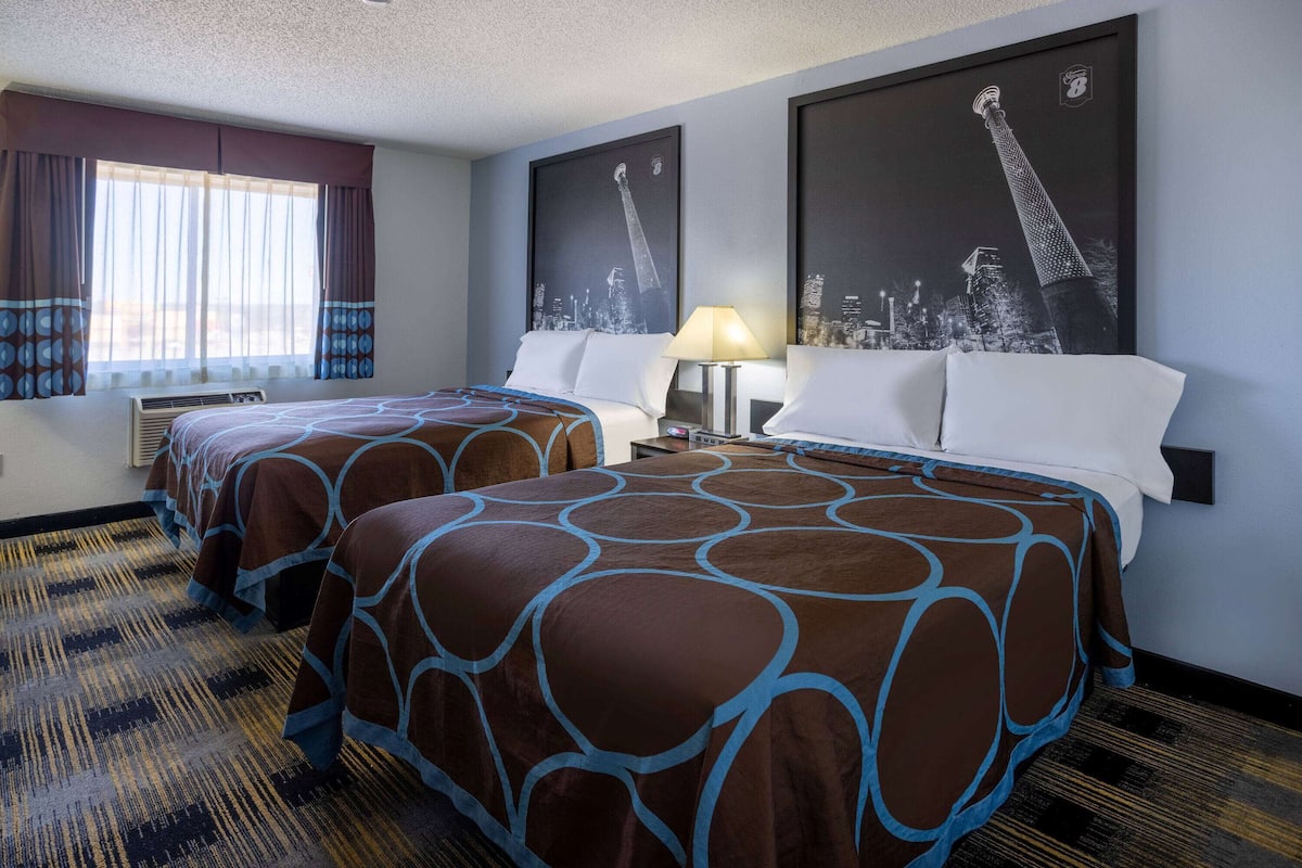 A hotel room with two beds, a large window with curtains, a cityscape artwork on the wall, and a patterned carpet.