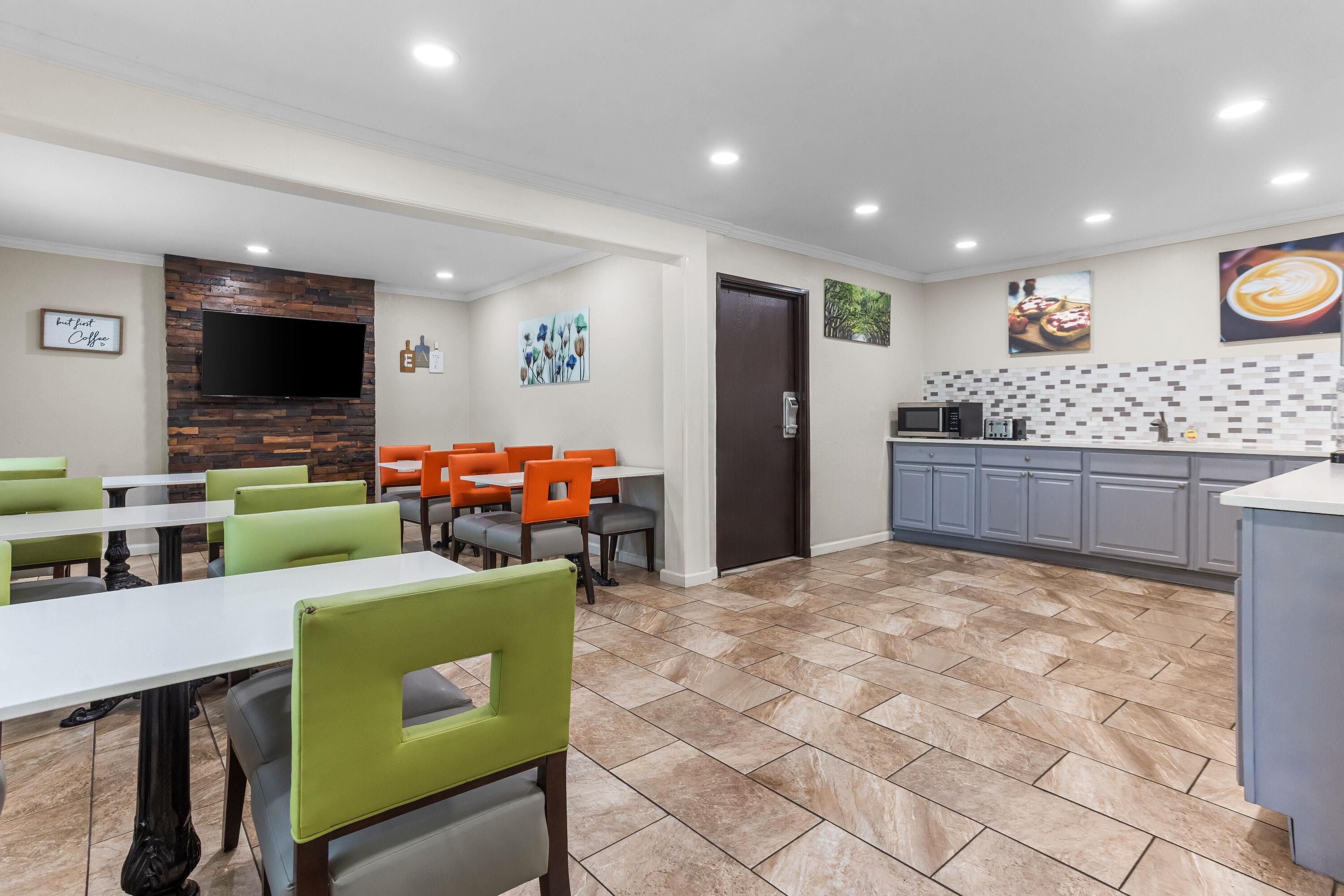 Un espace repas moderne avec des chaises vertes et orange, un téléviseur à écran plat fixé à un mur en bois, et une cuisine dotée de meubles gris et d’un dosseret carrelé.