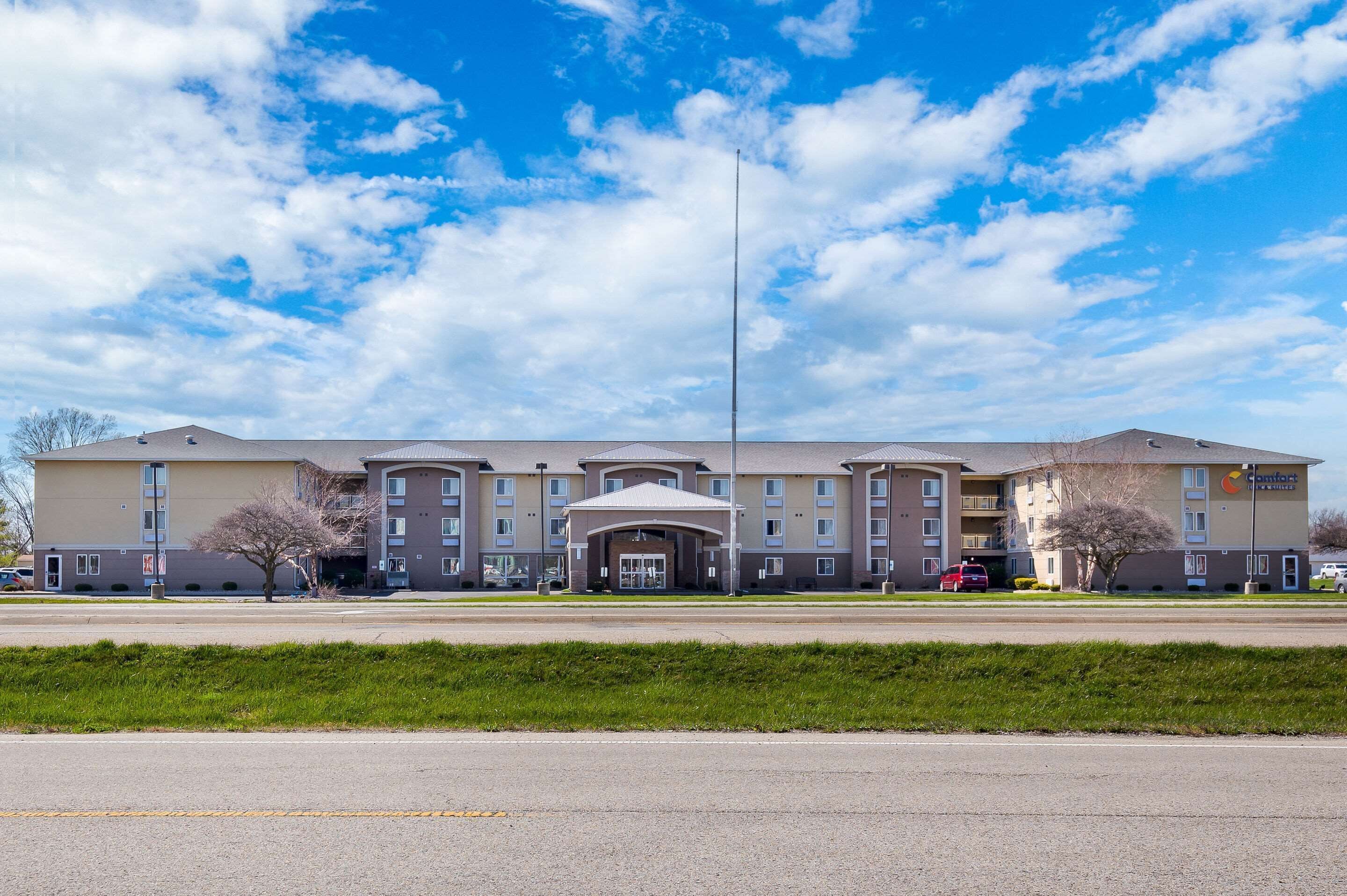 A multi-story hotel with a central entrance, parking lot, and a clear sky.