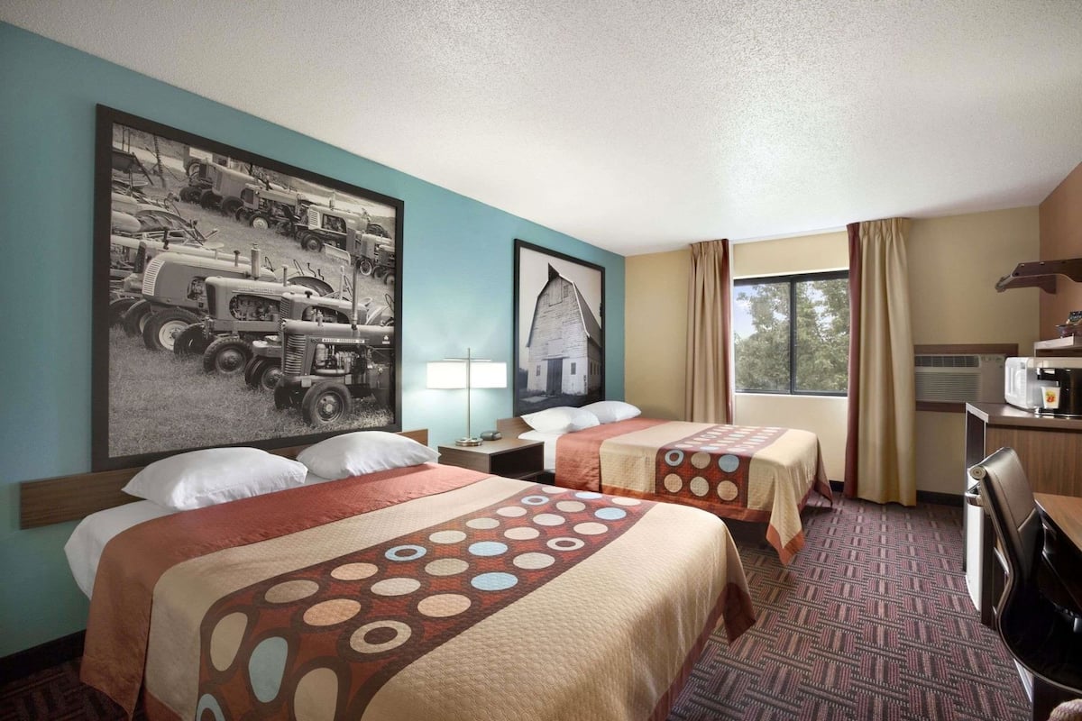 A hotel room with two beds, a desk with a microwave, a window with curtains, and a large wall painting.