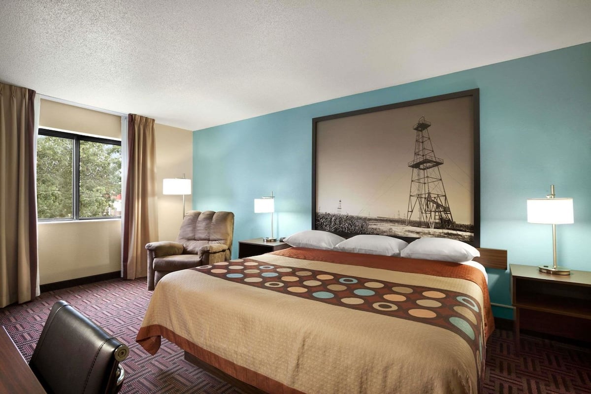 A hotel room with a bed, two bedside tables, a chair, a window with curtains, and a large wall painting.