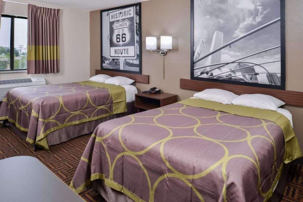 A hotel room with two beds, a window with curtains, a wall-mounted light, and a poster of a historic route.