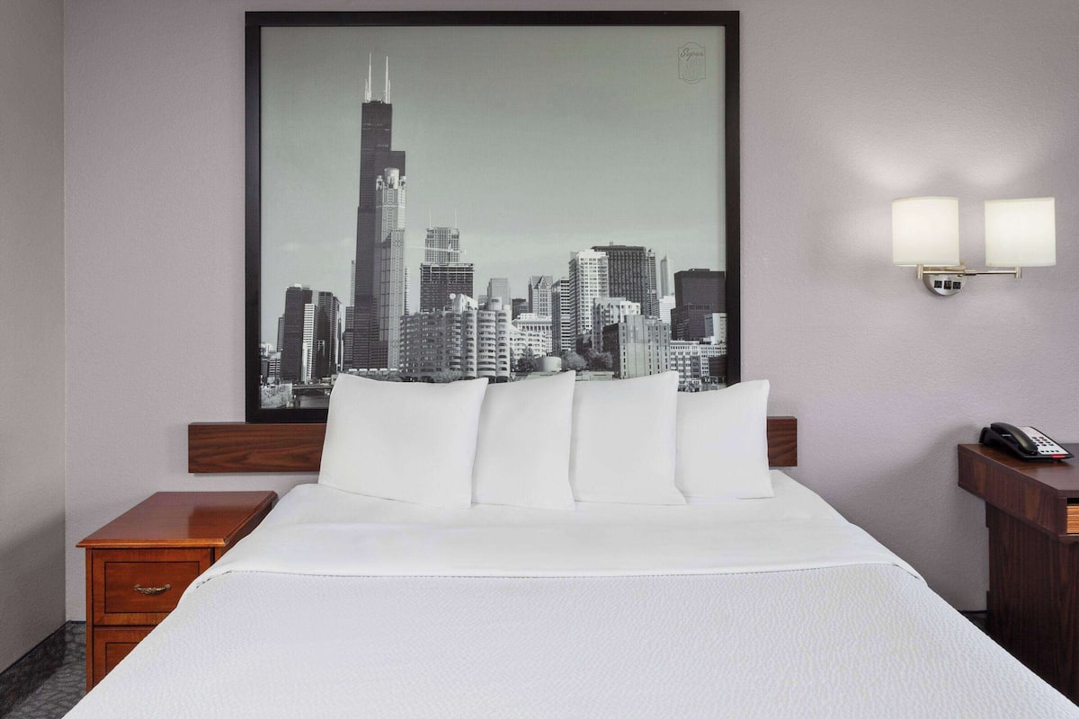 A hotel room with a large framed cityscape picture on the wall, a bed with white linens, a wooden nightstand, and a bedside table with a phone.