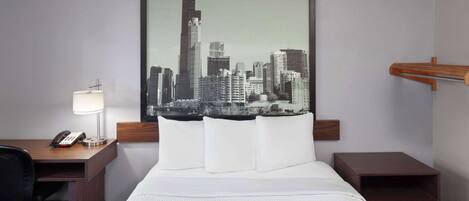 A hotel room with a bed, a desk with a lamp, a phone, and a cityscape painting on the wall.