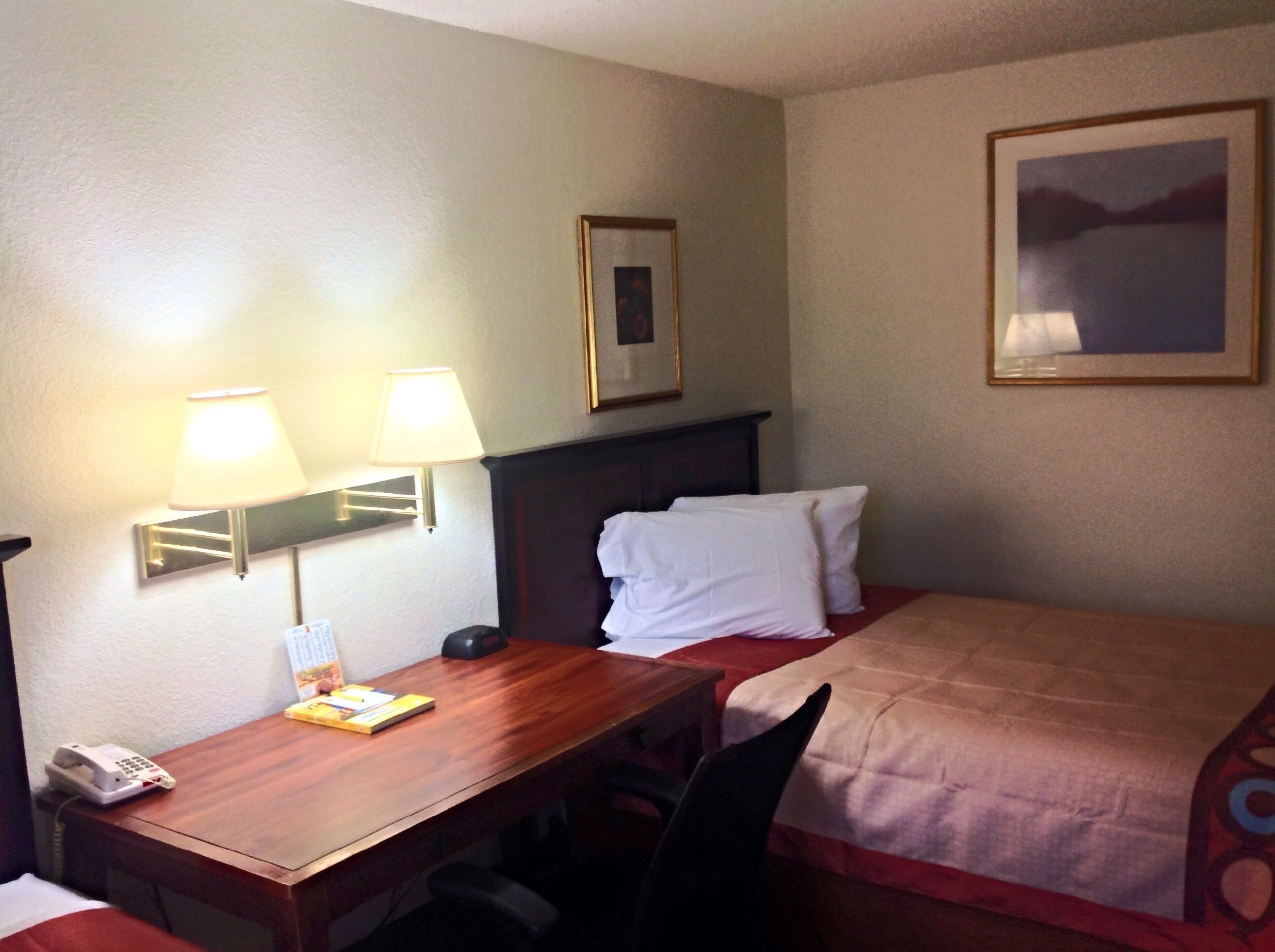 A hotel room with a wooden desk, two bedside lamps, a bed with white bedding, a framed picture on the wall, and a telephone on the desk.