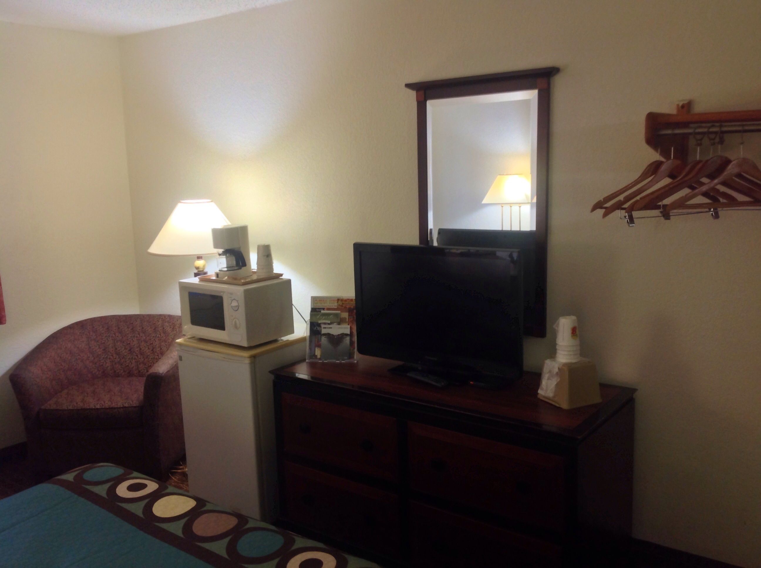 A hotel room with a bed, a desk with a computer and lamp, a television, a mirror, and a coat rack.