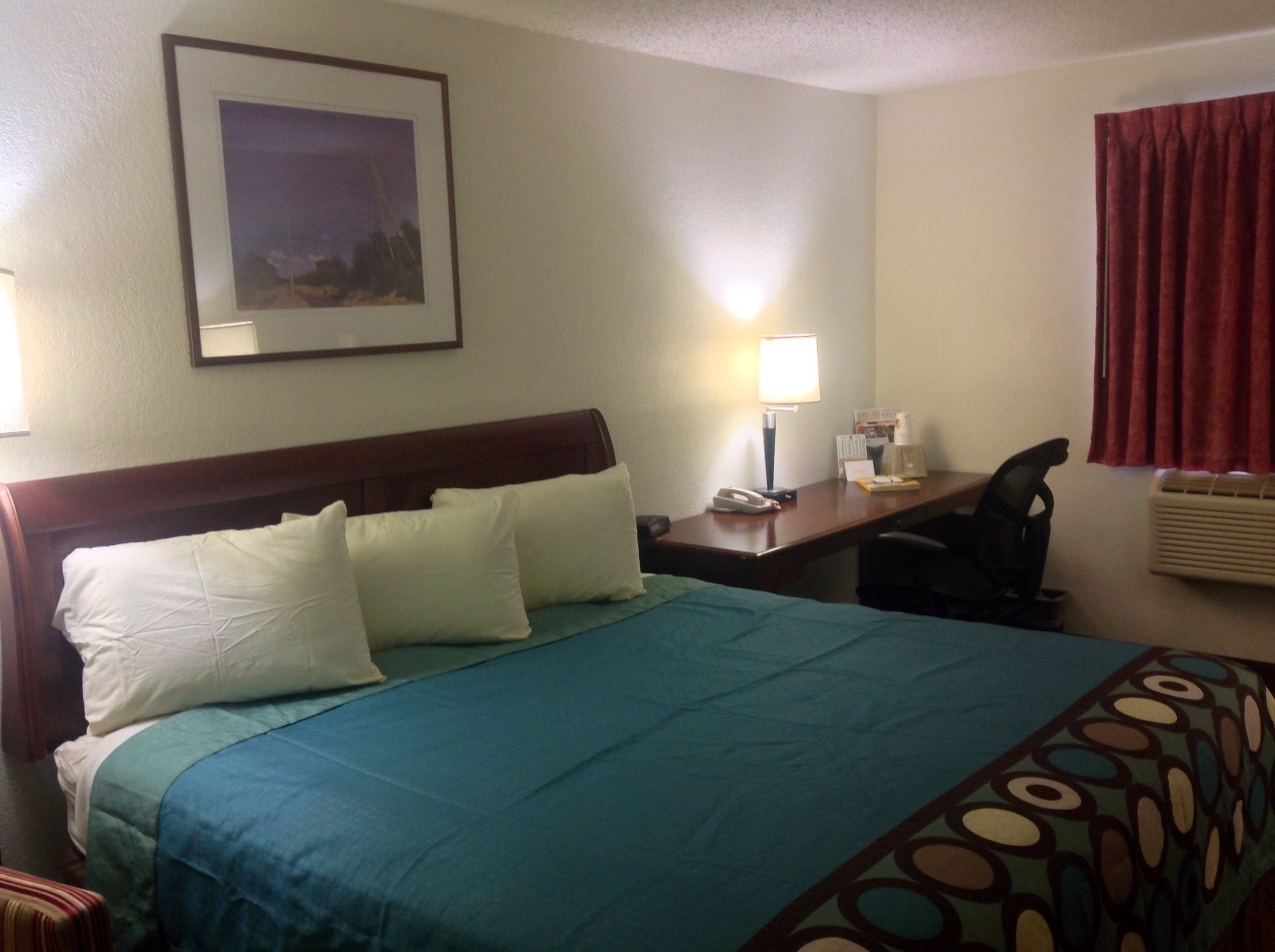 A hotel room with a bed, desk, chair, lamp, and a framed picture on the wall.