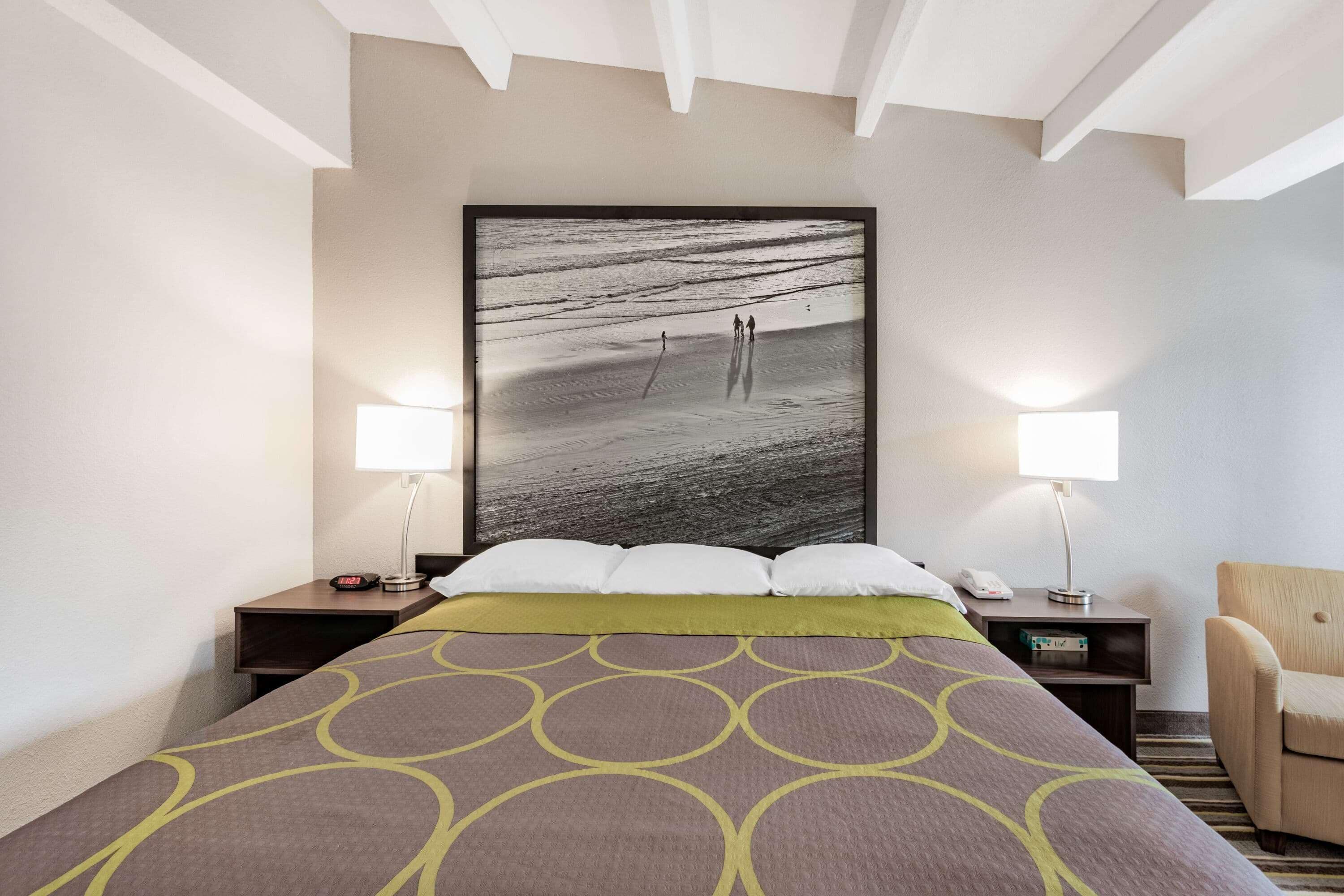 A hotel room with a large bed, two bedside tables, a chair, and a wall-mounted artwork of a beach scene.